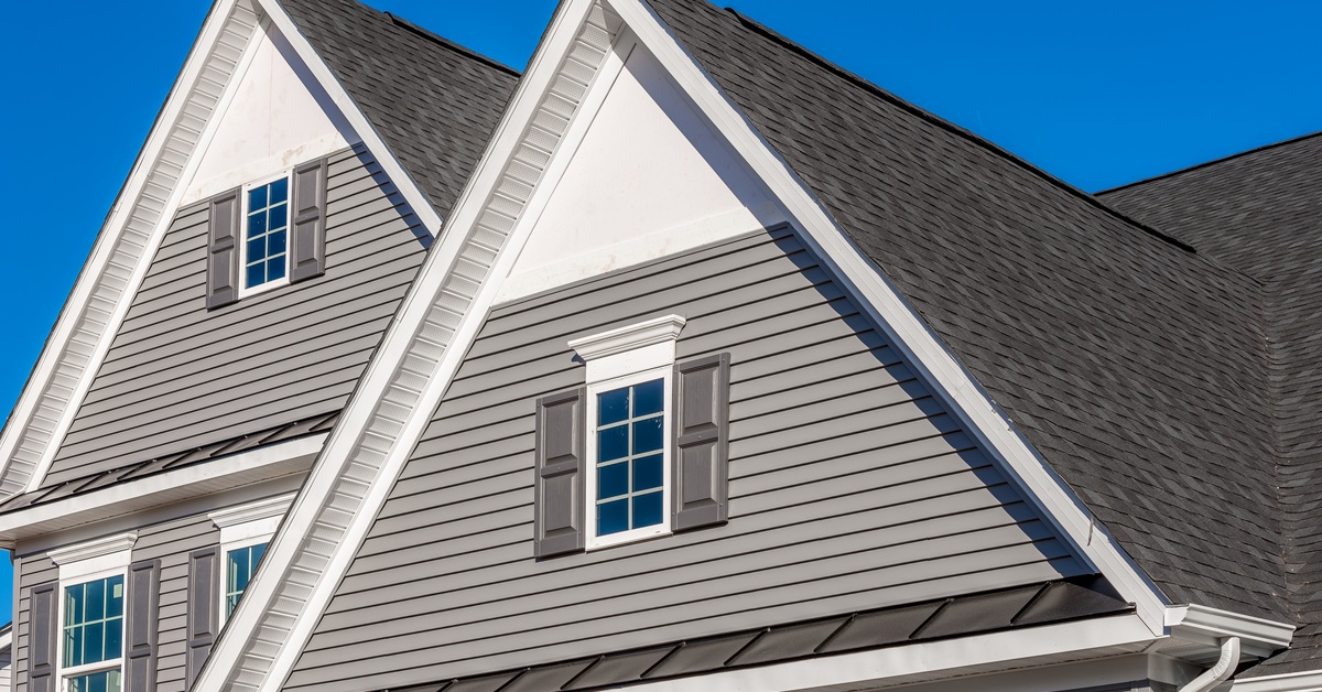 A close-up of a newly built home with white and gray decorative trim and gray vinyl siding on a sunny day.