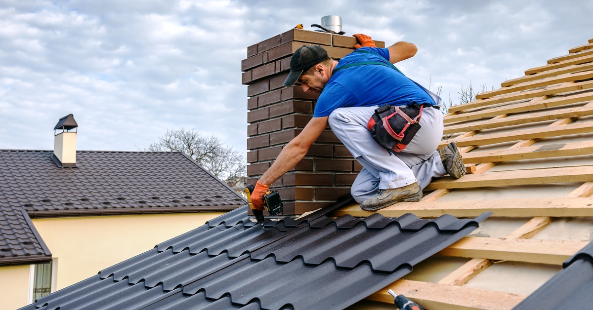 A worker wearing a blue shirt using a drill to attach black-colored metal shingles to the roof of a house.