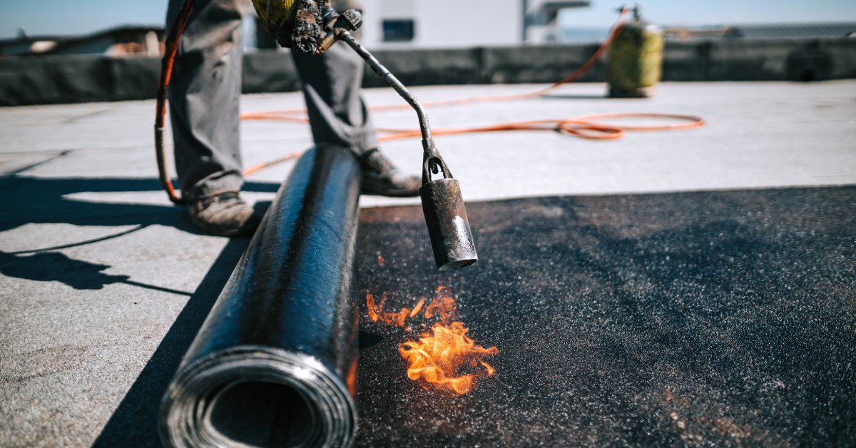 Roofing worker wearing a safety glove using a blowtorch to seal modified bitumen on the roof of a building.