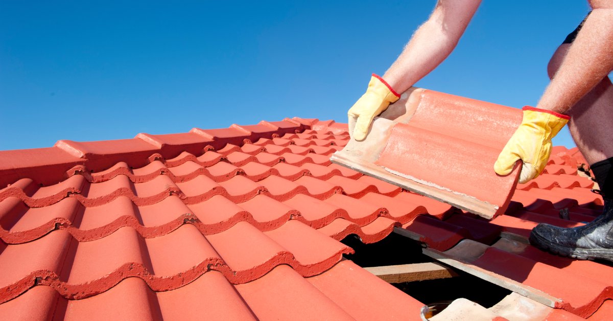 Roofing worker on top of a roof holding a red clay tile over a hole with multiple tiles laid all over the roof.