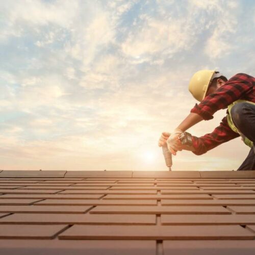 Worker wearing a plaid red shirt, yellow vest, and a helmet, using a pneumatic nail gun on a roof during sunset.