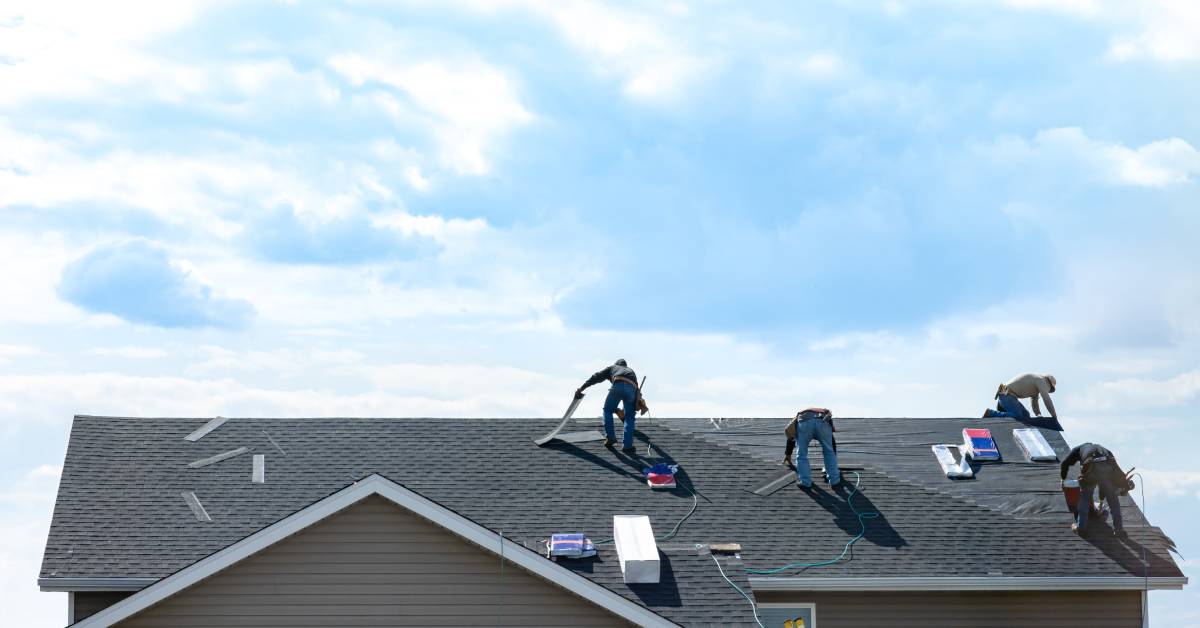 Four workers installing new grey shingles on the roof of a two-story home with a cloudy blue sky in the background.