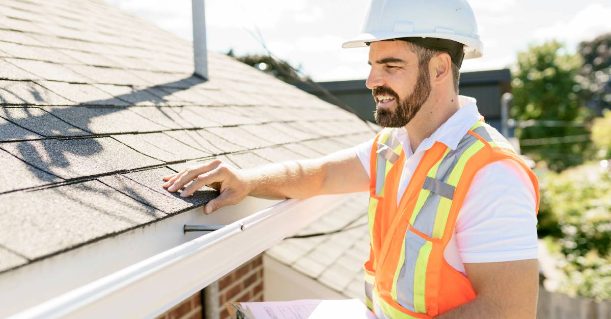 Man with a beard wearing a safety vest and a helmet carrying a clipboard checking the edge of a home’s roof.