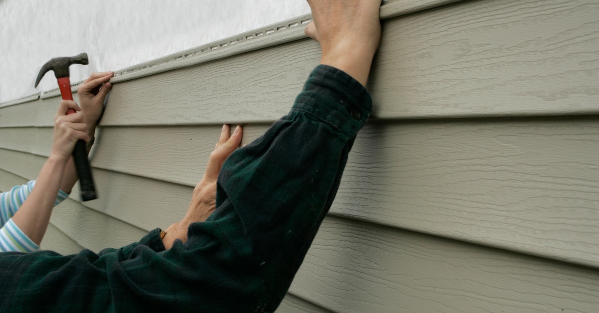 Two people are installing vinyl siding on a house. One holds up the siding while the other installs it using carpentry tools.