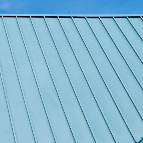 A close-up of a teal blue corrugated metal roof on a white home. The sky is clear with wisps of nearly transparent clouds.