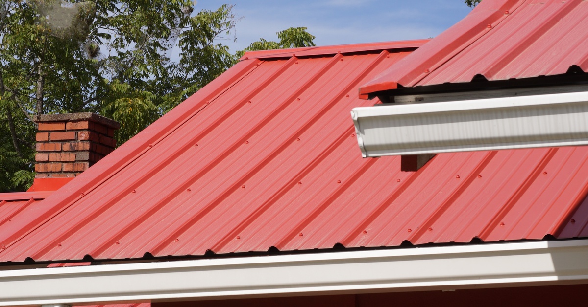 A close-up of a red metal roof with a red brick chimney on a residential building on a bright and sunny day.