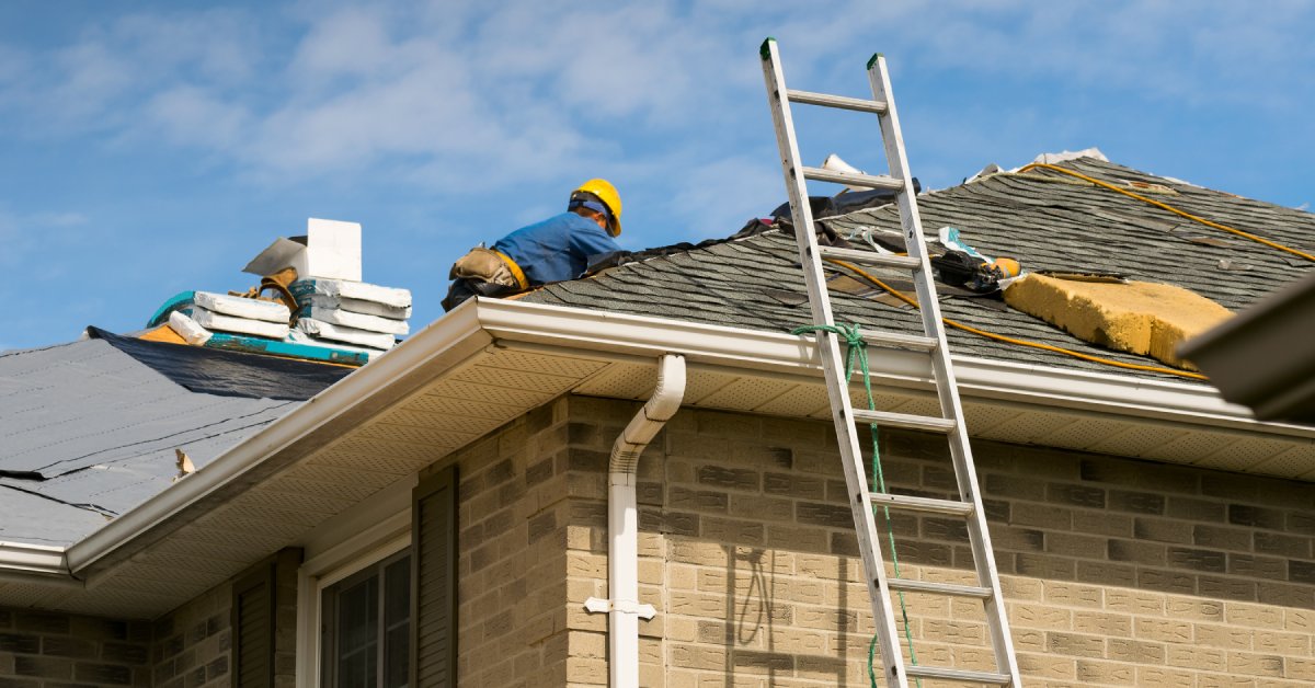 A roofer installing new shingles on the roof of an older house. They're wearing a yellow hard hat.
