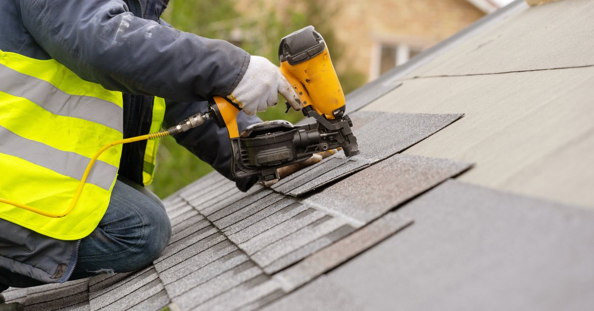 A close-up of a person using a nail gun to install tile on a roof. The person is wearing a yellow safety vest.