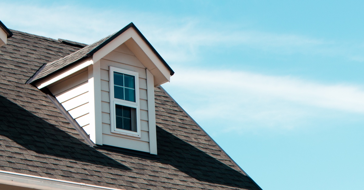 A roof of a classic home with brown shingles and a window with a white frame.