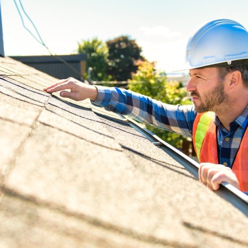 A man wearing an orange safety vest and a white helmet touching and looking closely at a shingle on the roof of a home.