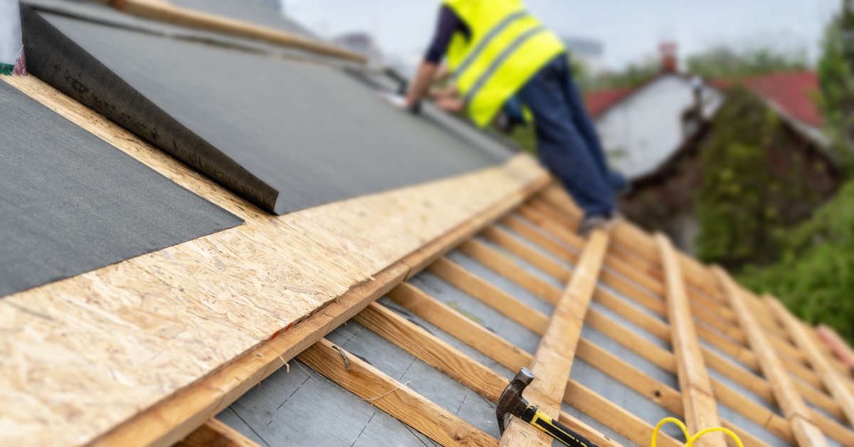 Construction worker standing on the structural wooden beam of a home’s roof while applying sheets of protection sheets.